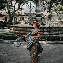 A traditional Mayan woman carrying textiles while walking in a park.