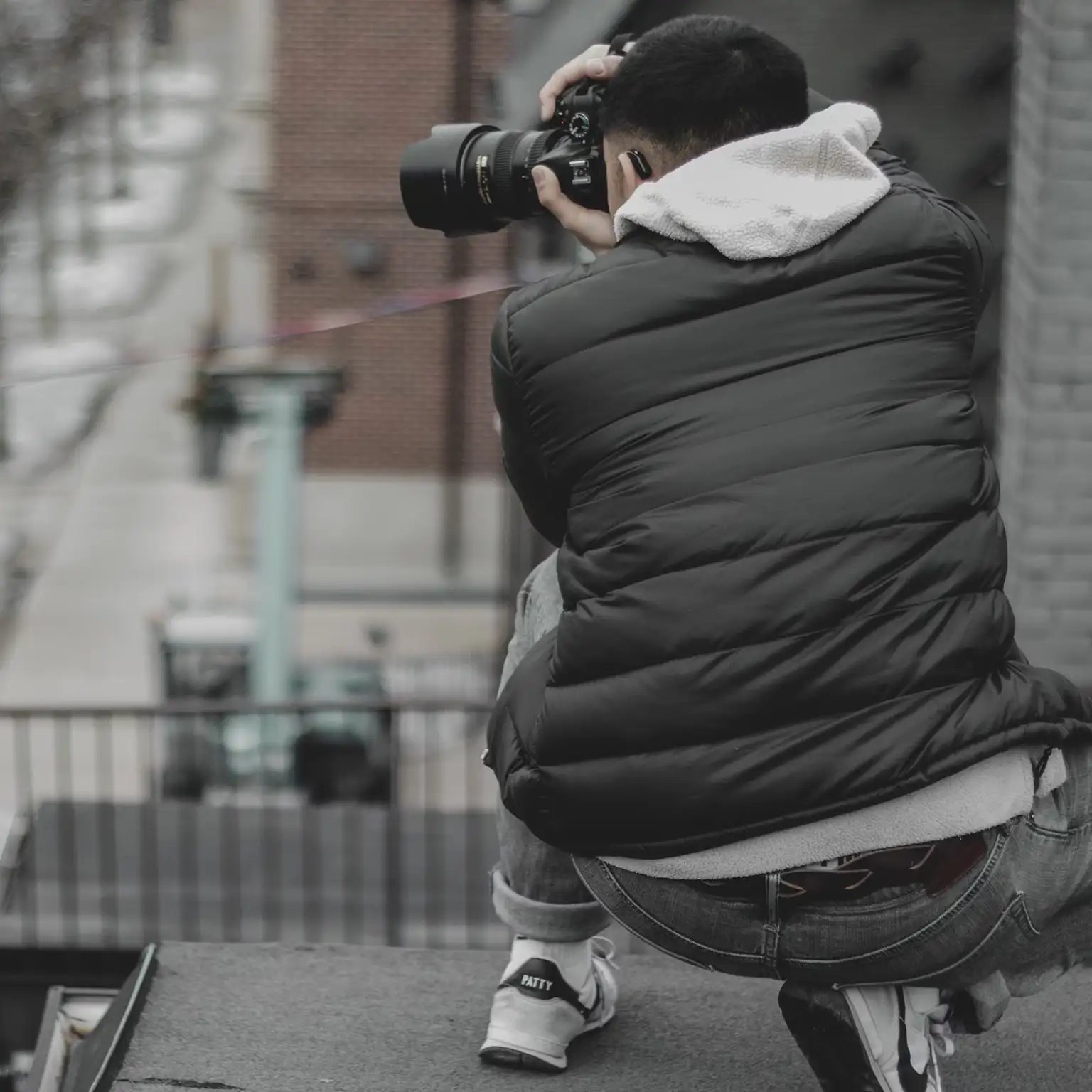 A photographer on a rooftop, dressed in a black puffer jacket, taking photos of the street below.