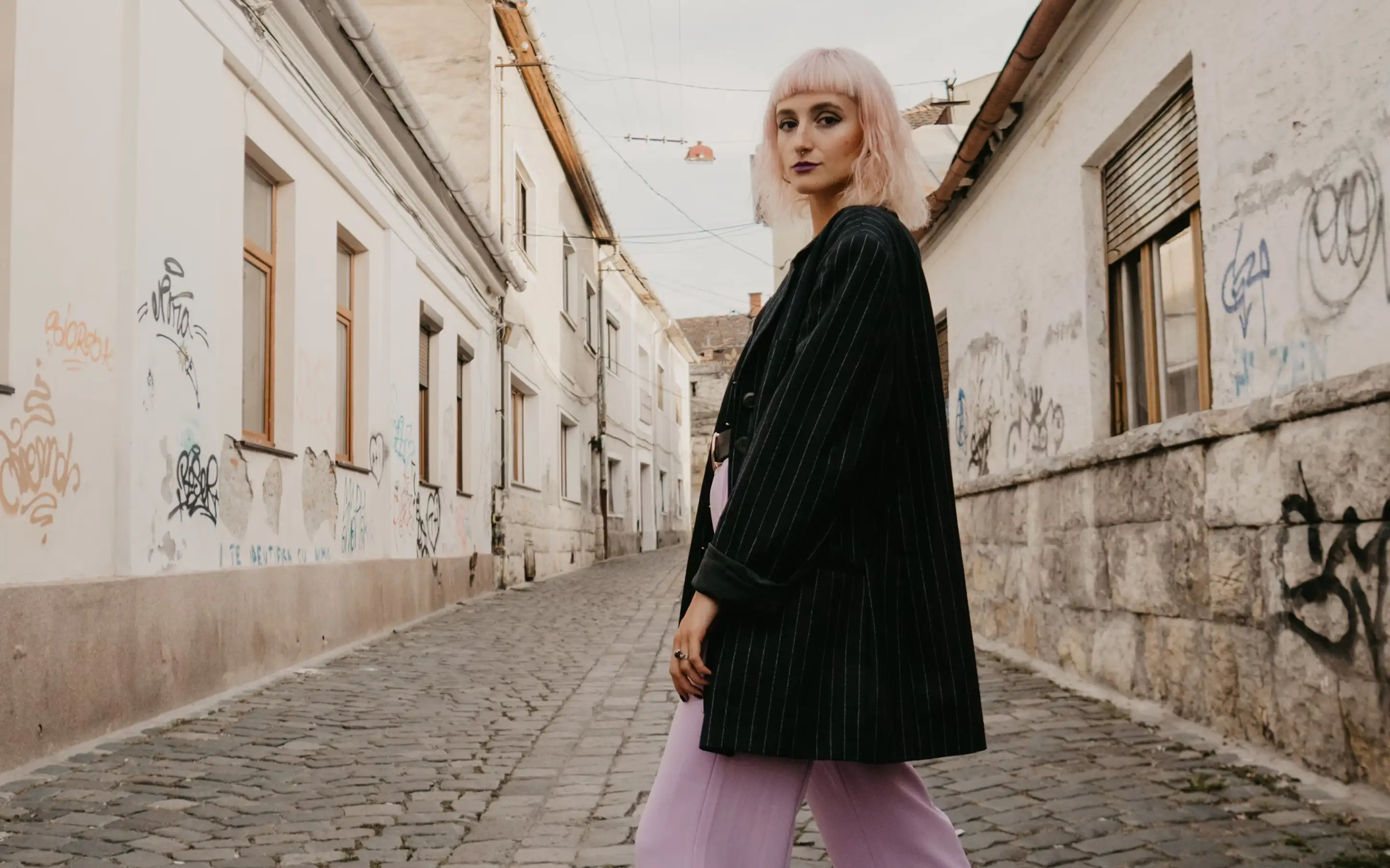 A trendy female model with pink hair posing in a stylish outfit on a city street.