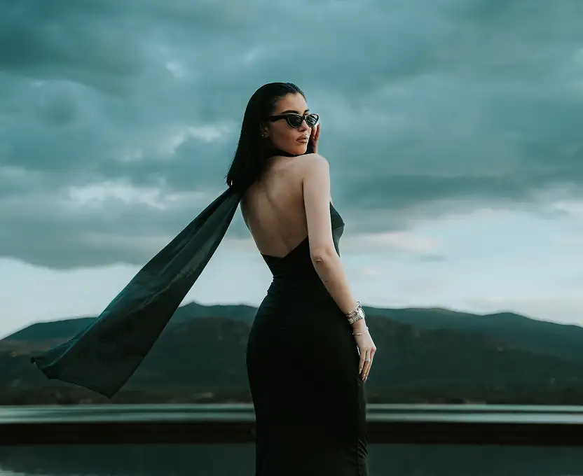 Elegant woman wearing a black dress with a flowing scarf against a dramatic sky.